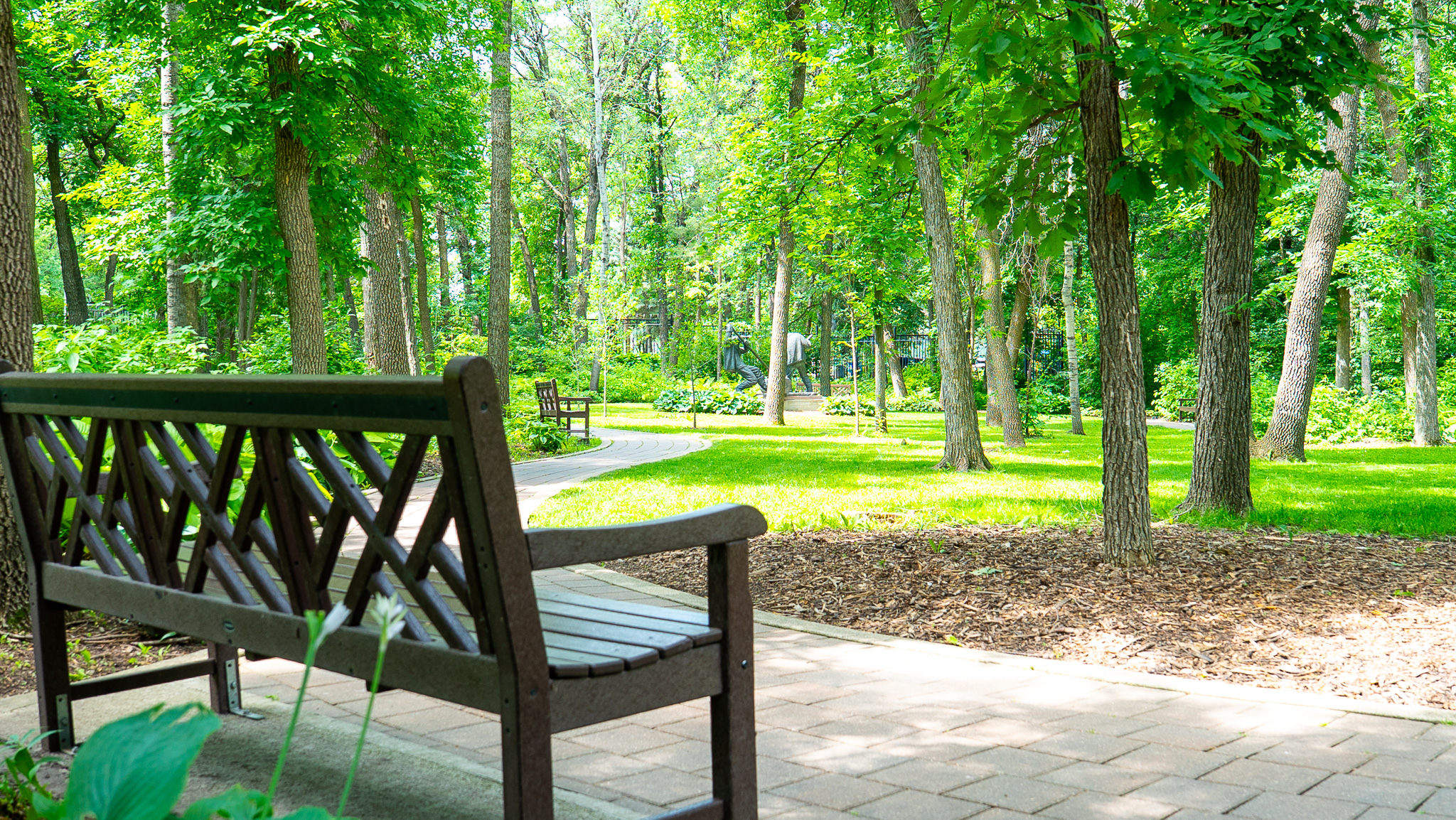 Shaded bench and brick pathway beside sculptures in the Leo Mol Sculpture Garden at Assiniboine Park