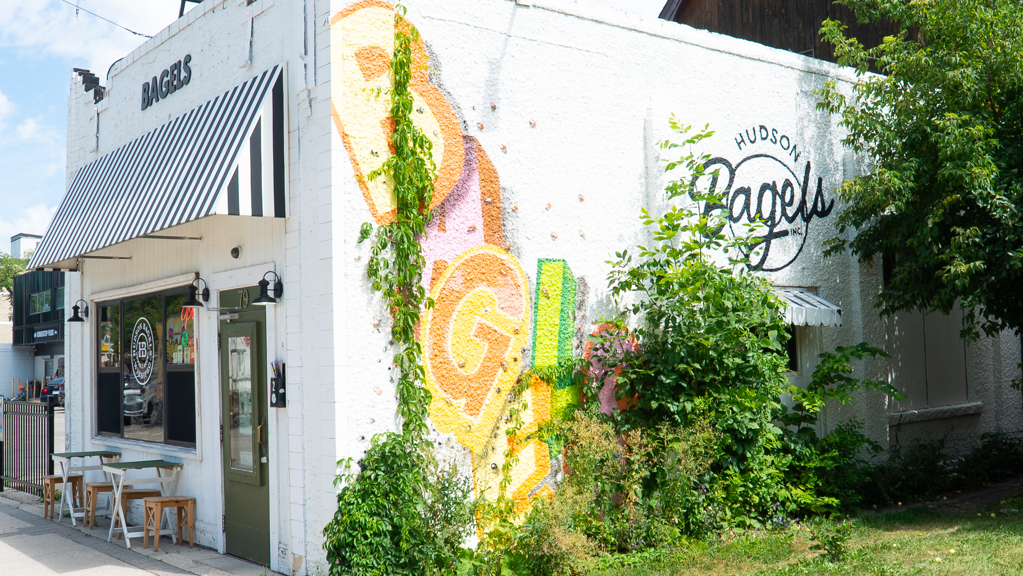 White brick exterior of Hudson Bagels with a colourful painted mural and striped awning