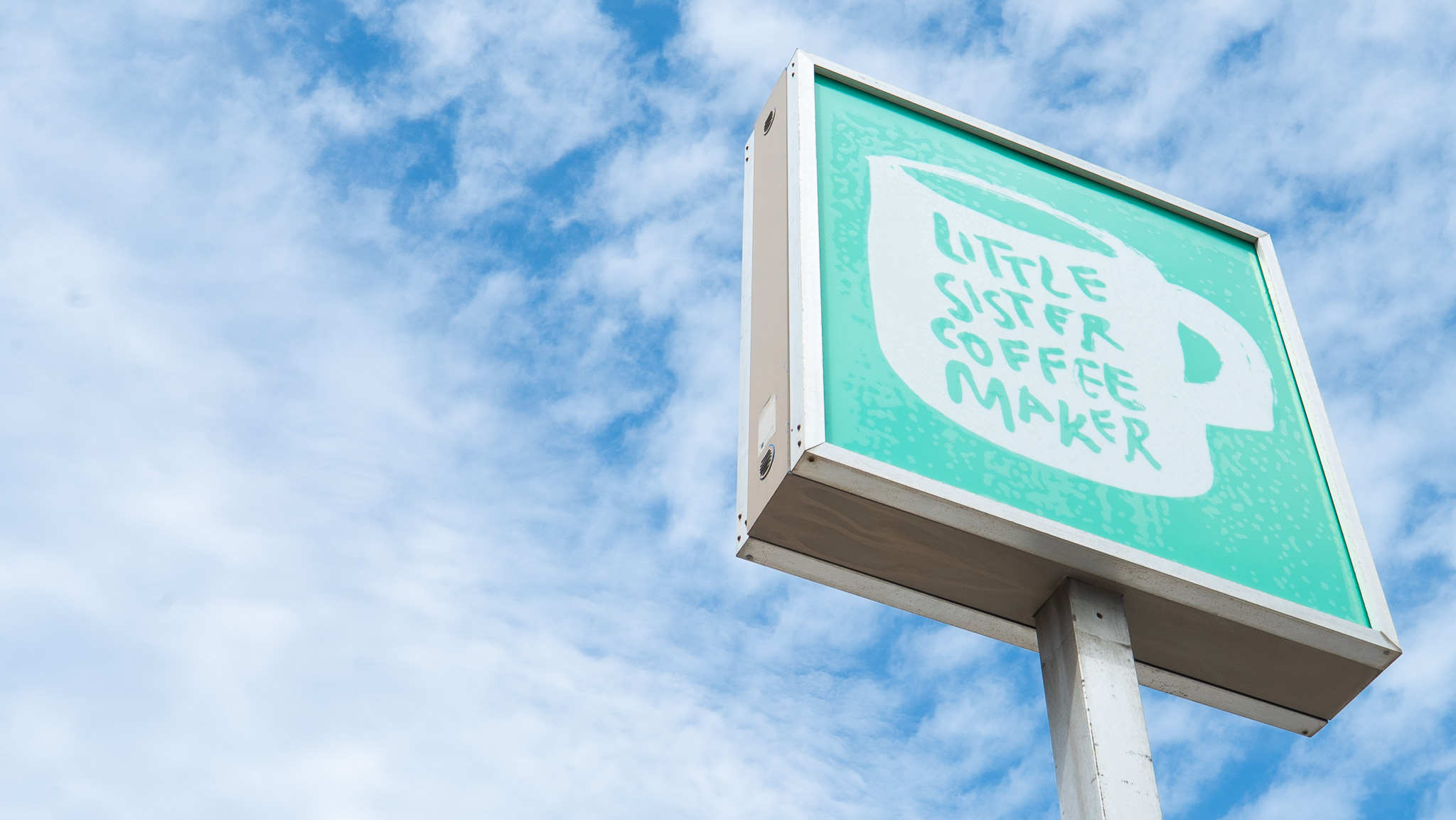 Little Sister Coffee Maker sign with a mint green coffee cup logo against a blue sky